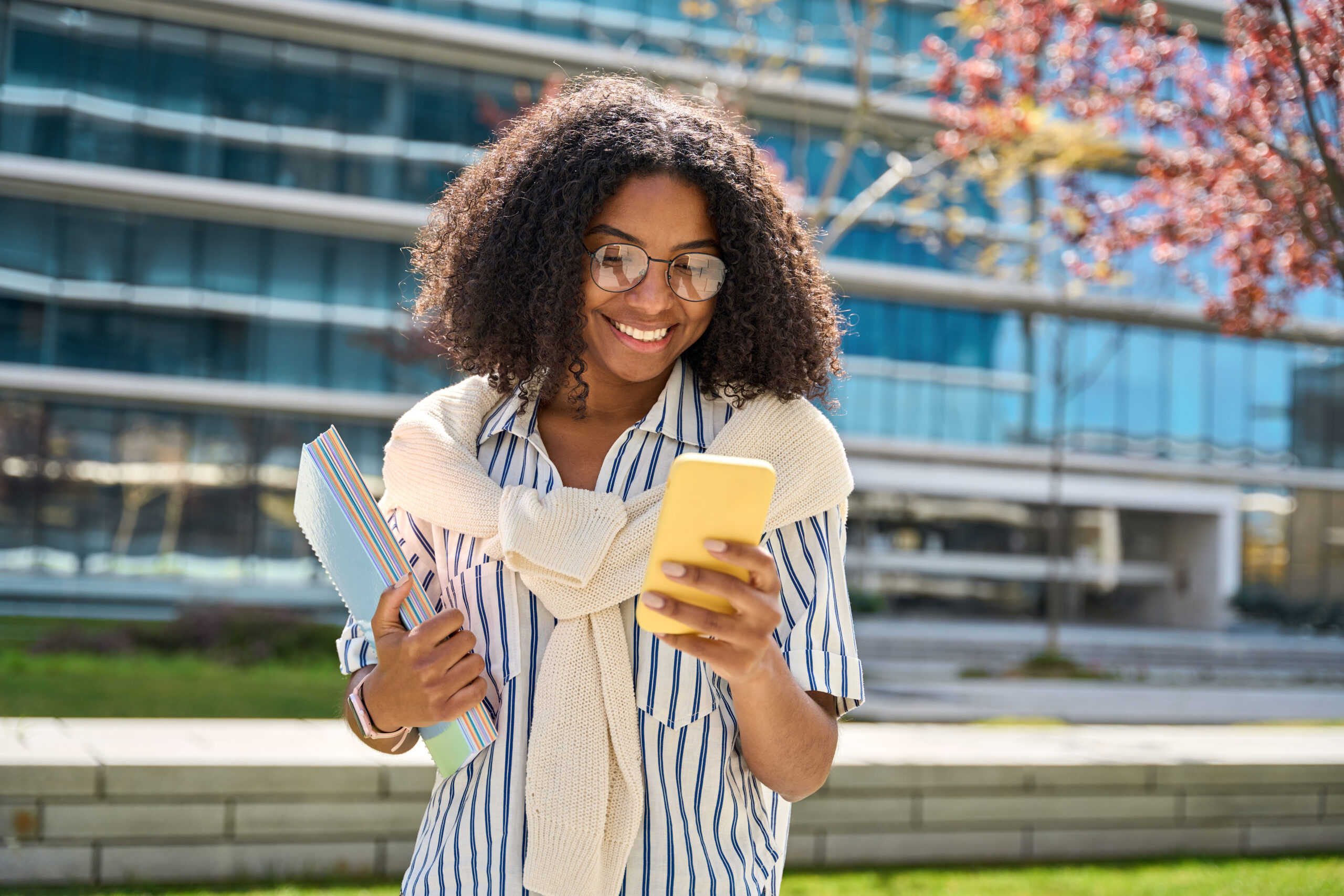 Young happy African American girl walking in city park using mobile phone shopping online or texting messages. Smiling female student holding cellphone chatting going along campus area.
