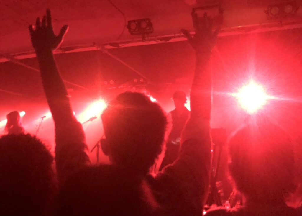 Photo of crowd at Mount Kimbie. Red stage lighting. Person with hands in the air.