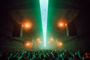 Image of crowd at Warehouse Project - dancing to the DJ whilst a beam of green light dramatically shines over the crowd from the stage. It is a big room full of people.
