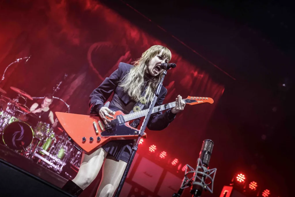 Lead singer of Halestorm at the AO Arena, Manchester. She is singing passionately into the microphoene, playing a red electric guitar. The backdrop of the stage is lit in red light.