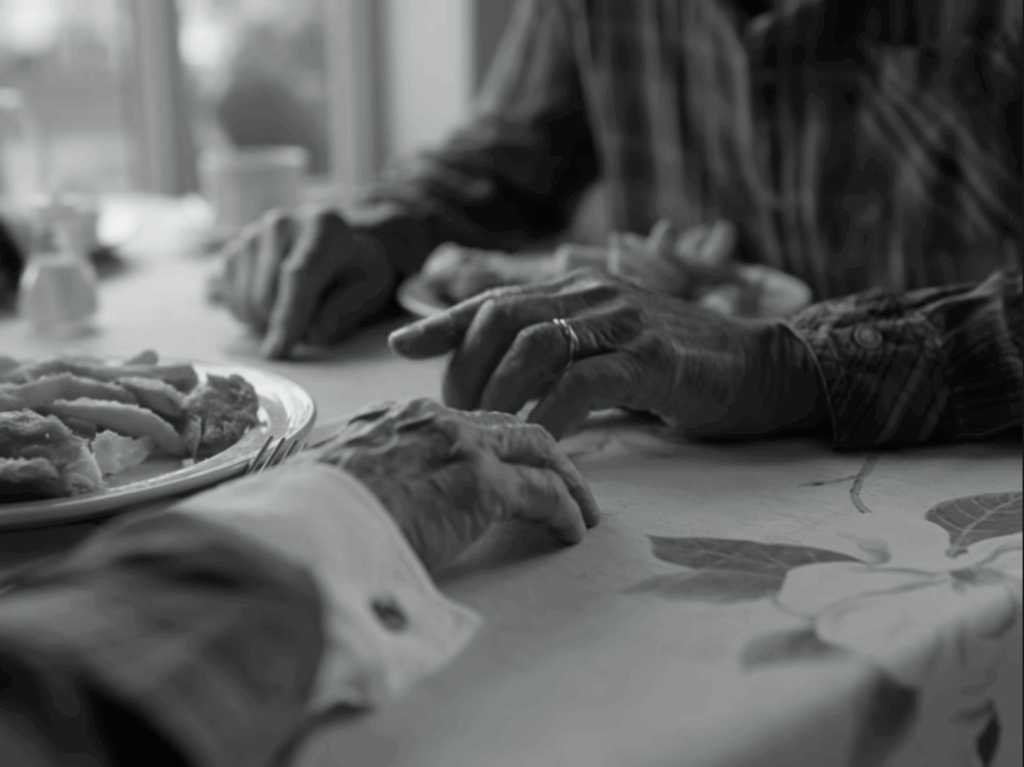 An older couple sitting at a table in a fish and chips shop. The photo is a black and white image of just their hands on the table, close to each others, next to the plate of fish and chips. It has a tender atmosphere.