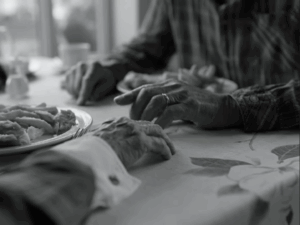 An older couple sitting at a table in a fish and chips shop. The photo is a black and white image of just their hands on the table, close to each others, next to the plate of fish and chips. It has a tender atmosphere.