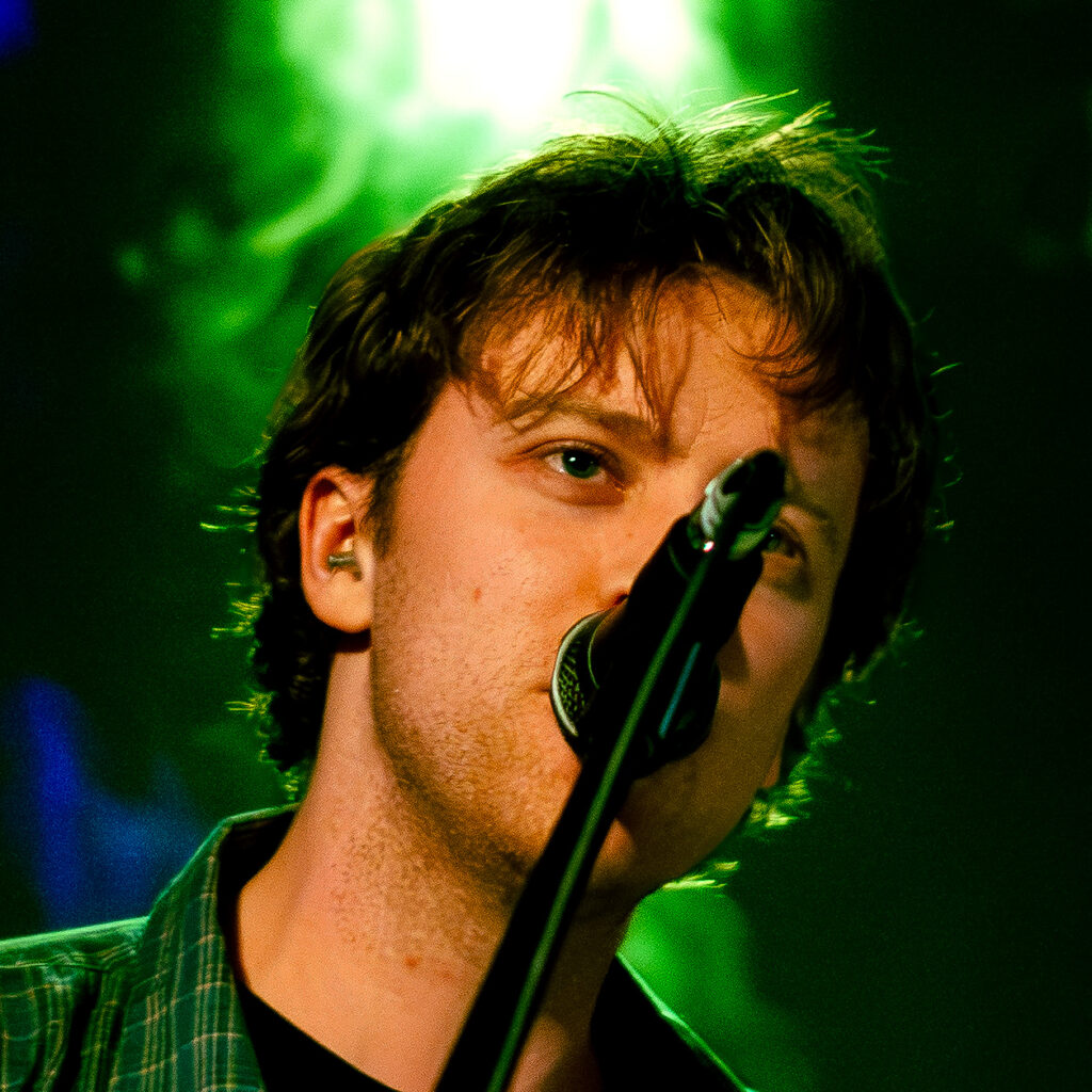 Close-up shot of singer and guitarist, James Bradbury, singing into the mic which partially covers his face. The stage lighting in the background is an atmospheric dark green.