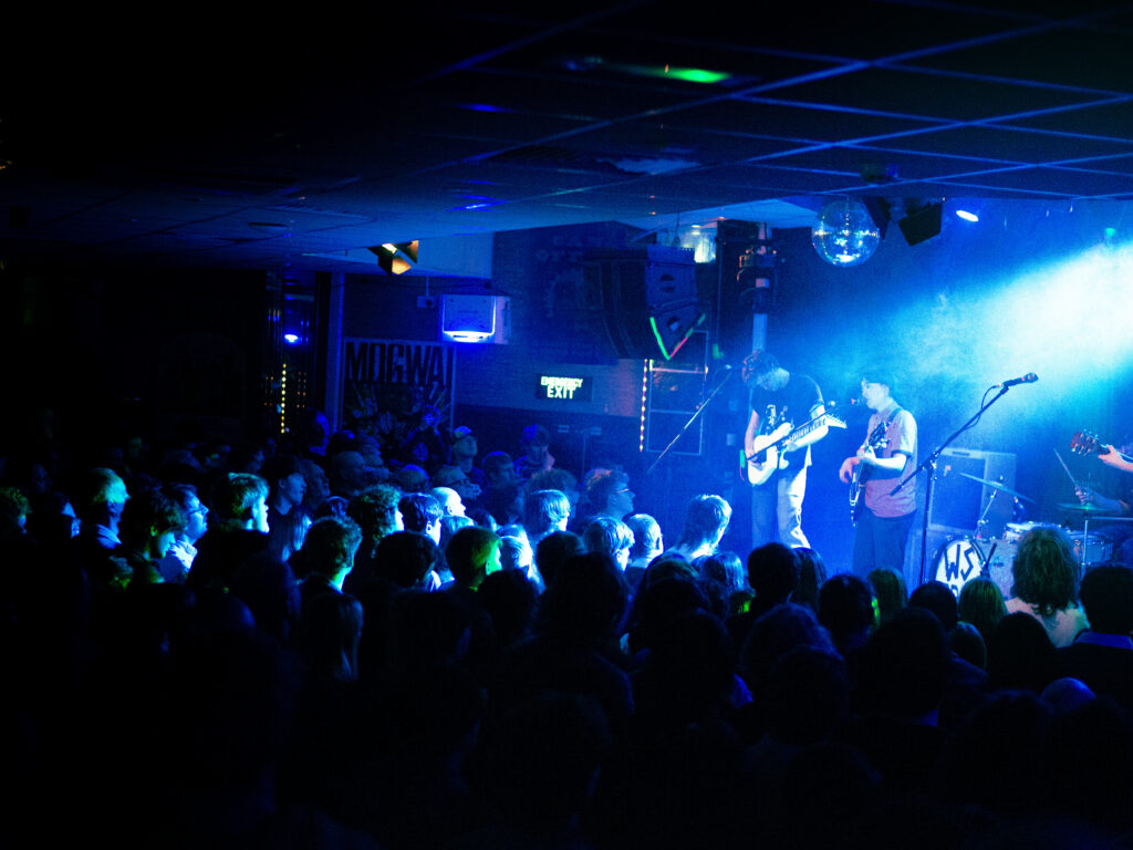 Wide shot of the crowd and the band onstage taken from the back of the crowd. The room is dark except for the vibrant blue stage lights.
