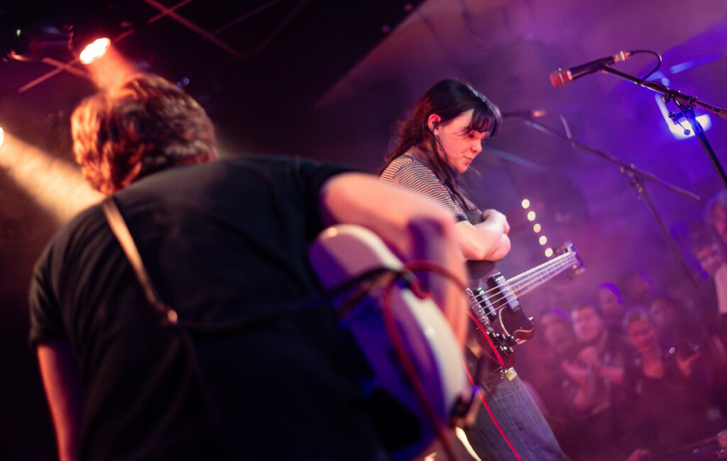 Wide shot of Westside Cowboy on stage taken from the side of the stage.  There is the back of the guitarist in the foreground and further onto the stage, Aoife (bass guitar and vocals) is getting ready to sing. Part of the crowd can be seen in the bottom corner, also bathed in the warm purple glow of the stage lighting.