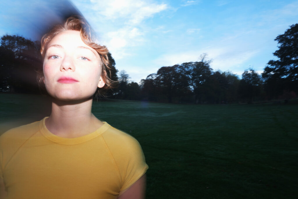 picture of Neve Cariad in a field, trees in the background. the sky is blue and the lighting is arty in style - overexposed on Neve's face. She is wearing a yellow top.