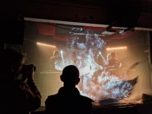 Picture of the stage taken from the crowd. There is a mesh gauze in front of the band which has abstract images projected onto it, creating an ethereal effect.