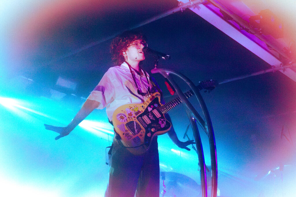 Cavetown onstage, singing into the mic with guitar slung on - arms outsretched either side. The photography style has played on their colourful style with rainbow-esque lights surrounding the image.