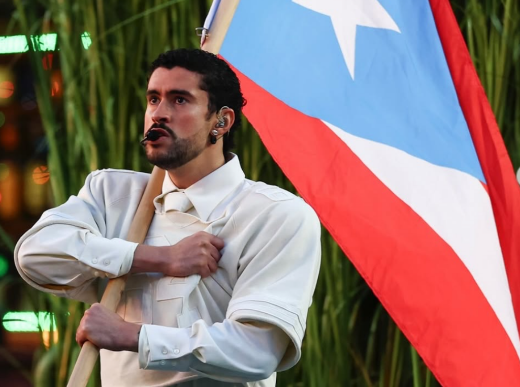 Bad Bunny performing at the Superbowl Half-Time Show in 2026. He has a serious or concentrating expression and is holding a large flag of Puert Rico.