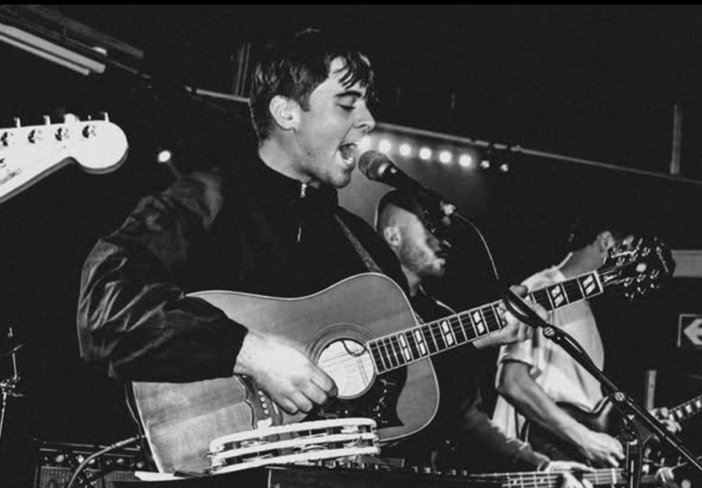 Musician from band 'The Cavs' playing acoustic guitar and singing onstage. Black and white photo