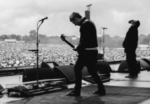 Black and white landscape image of Noel Gallagher performing onstage at a festival.