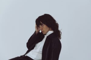 Press photograph of Courtney Barnett, leaning back on chair dressed in a black suit jacket and trousers and white shirt.