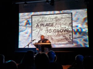photo of Warrington-Runcorn New Town Development Plan performing onstage in front of a screen that reads 'A PLACE TO GROW'.