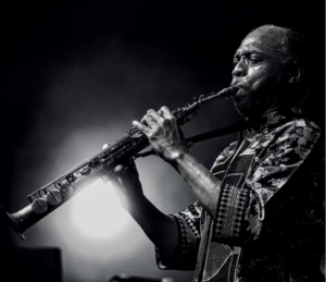 Black and white photo of Femi Kuti performing live. Dramatic lighting make it an impressive shot of him mid-playing a wind instrument