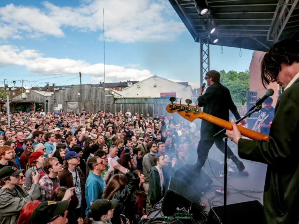 wide shot of band Yard Act onstage at temporary stage in Brudenell car park. The sky is blue with few clouds, the crowd is full and colourful and attentive to the stage. James Smith, the frontman, is mid-performance. At the foreground, to the side, you can see the guitarist and his guitar.