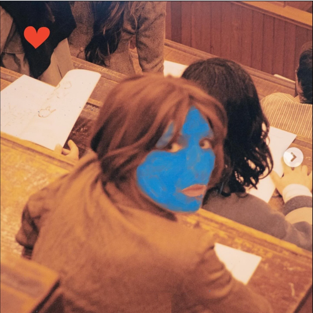 Album cover for Aldous Harding's 2026 release 'Train on the Island'. It features a photo of Aldous in what looks like a lecture theatre - her face is turned to face the viewer, but the photograph has been drawn over so that her face is bright, light blue. There is a small red love heart in the top left corner.