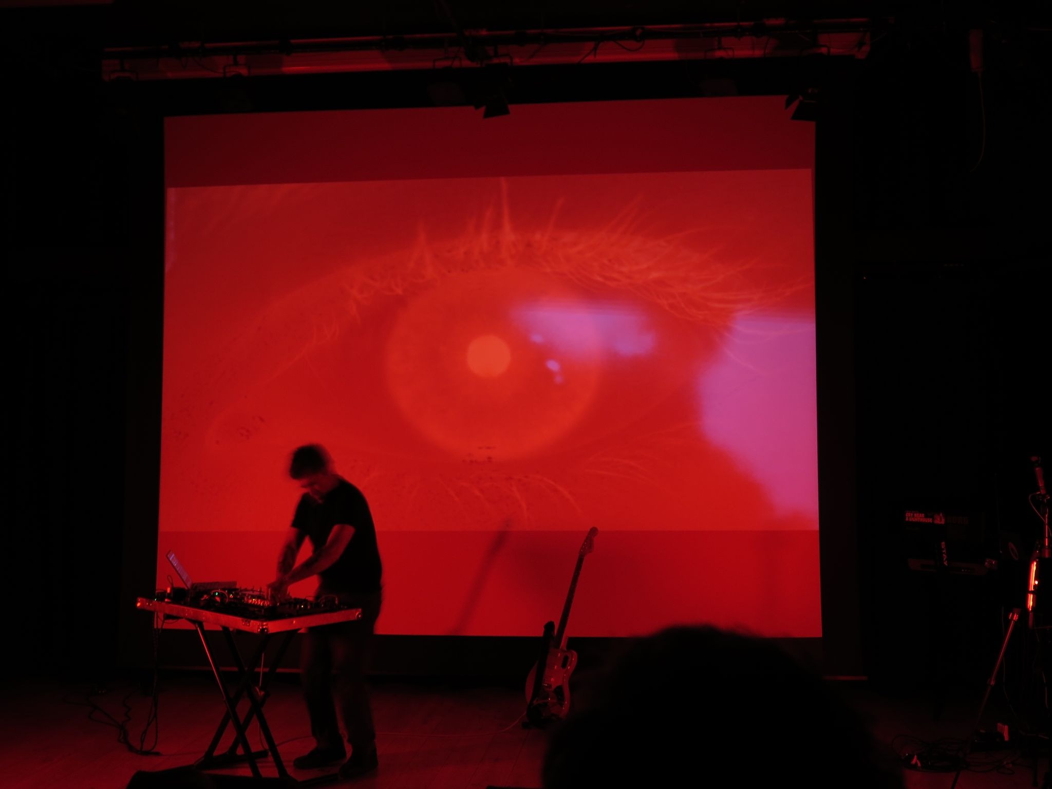 A picture of Thomas Ragdale (a.k.a. Sulk Rooms) playing a synth keyboard in front of a projector screen with a red-tinted image of an eye.