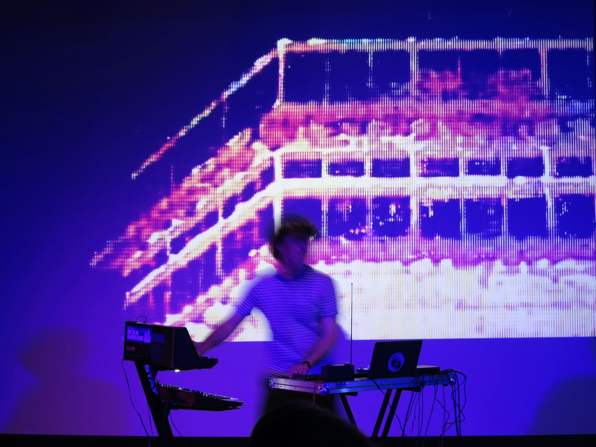 Picture of James Adrian Brown playing a synth deck in front of a blue-tinted projecter screen, his figure blurry as he moves from one to the other.